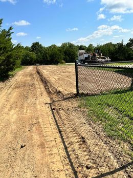 Dirt road beside a chain-link fence under a sunny blue sky with trees and construction equipment in the distance