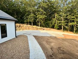 Curved concrete walkway beside a white building, leading to a gravel and dirt yard with trees beyond