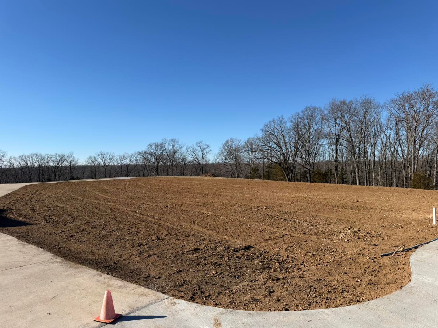 Freshly graded dirt lot beside a paved road under a clear blue sky, with trees in the background.