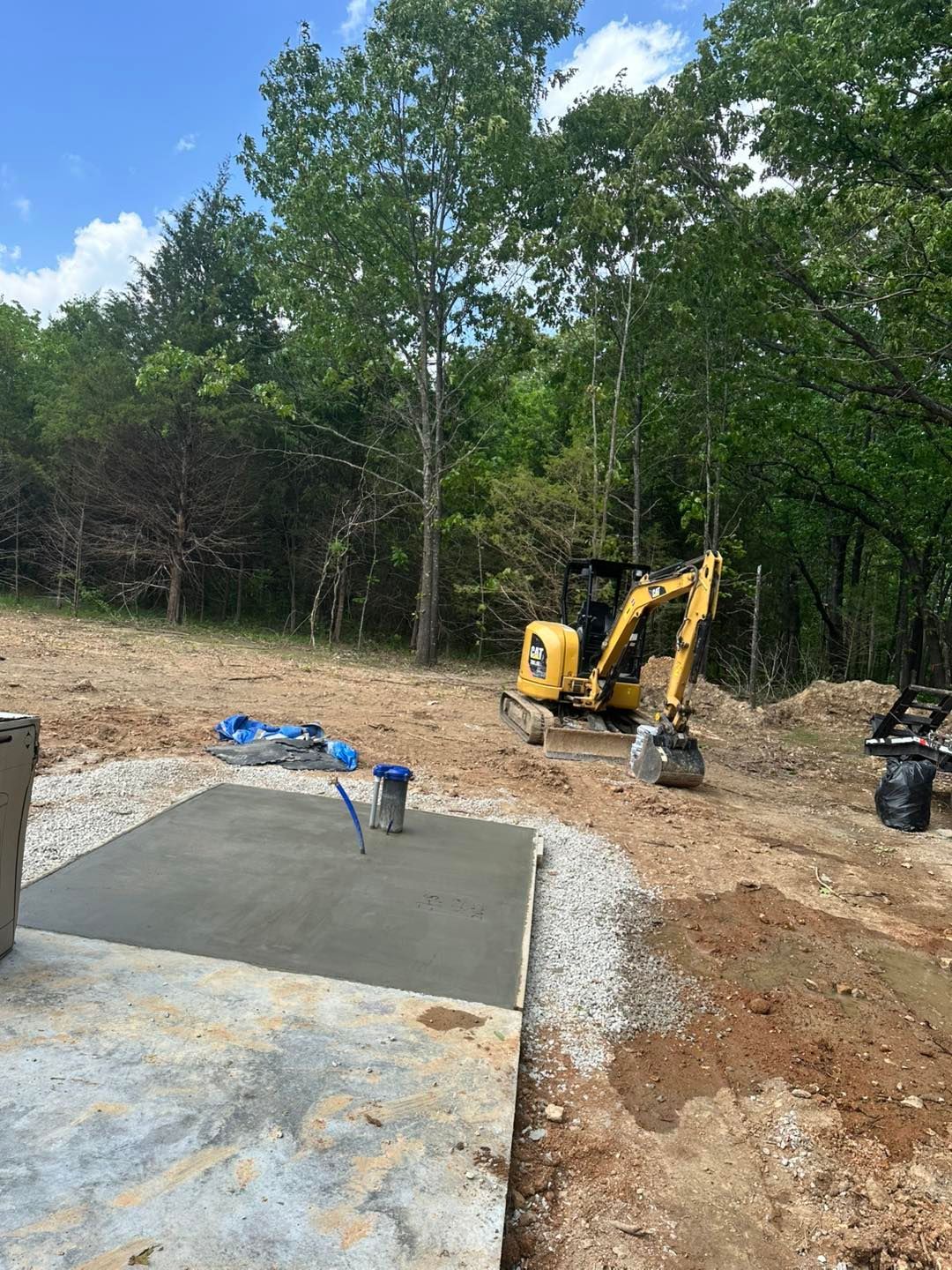 Backyard construction site with a yellow excavator beside a freshly poured concrete slab.