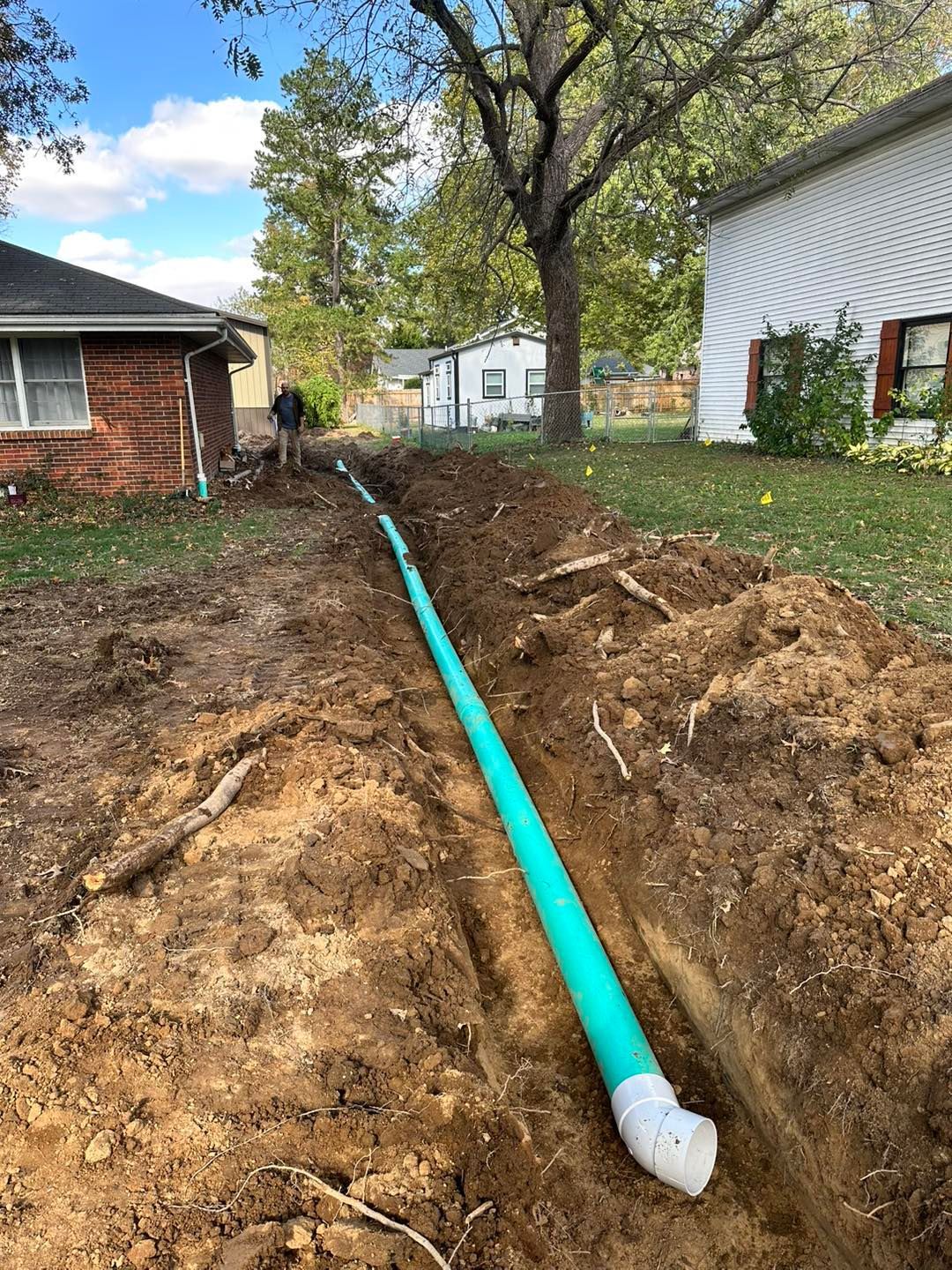 Trenched yard with a bright blue pipe running between houses, surrounded by dirt and trees.