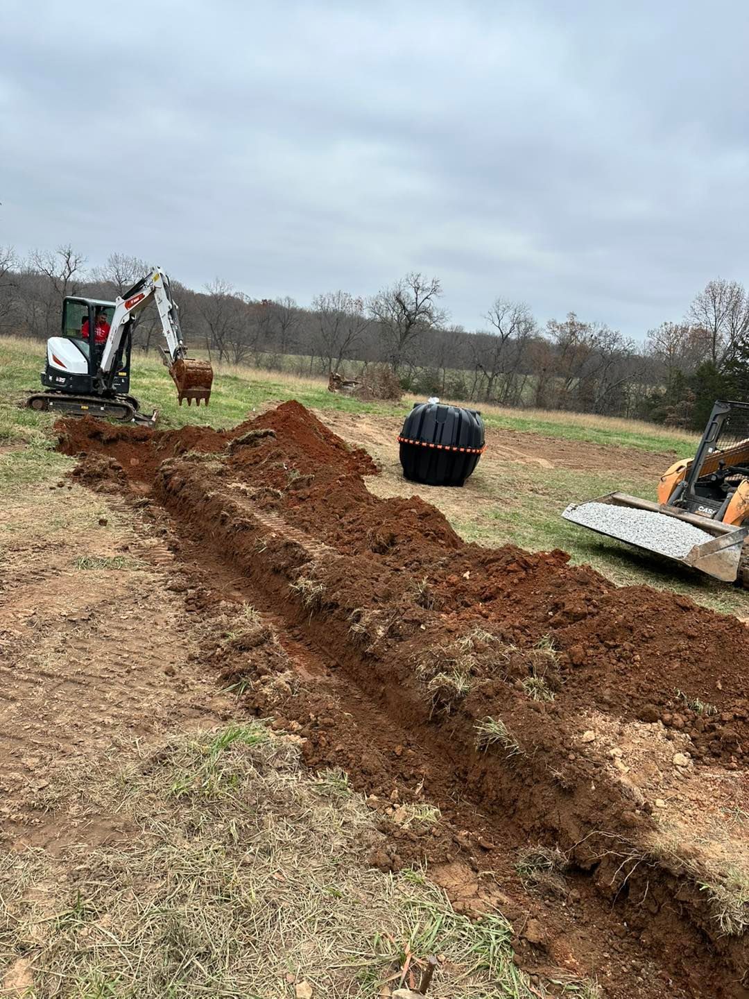 Excavator digging a long trench in a muddy field under cloudy skies, with a wheelbarrow and dirt piles nearby