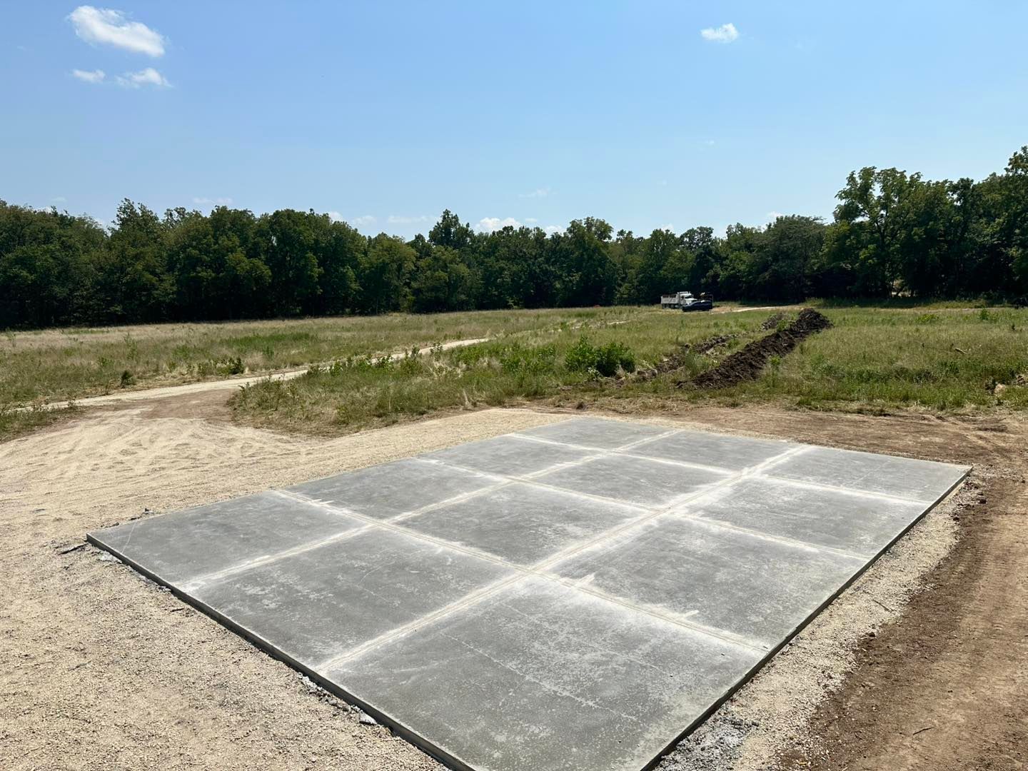 Concrete slab in a gravel lot beside a dirt path, with grassy field and trees under a blue sky