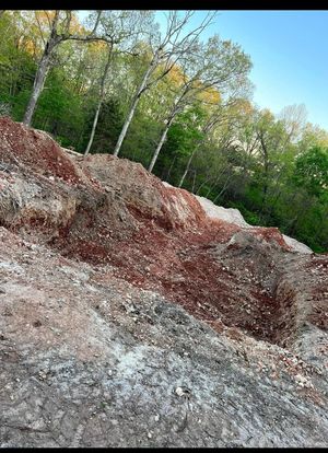 Rocky dirt embankment with red and gray soil beneath leafy green trees and a blue sky