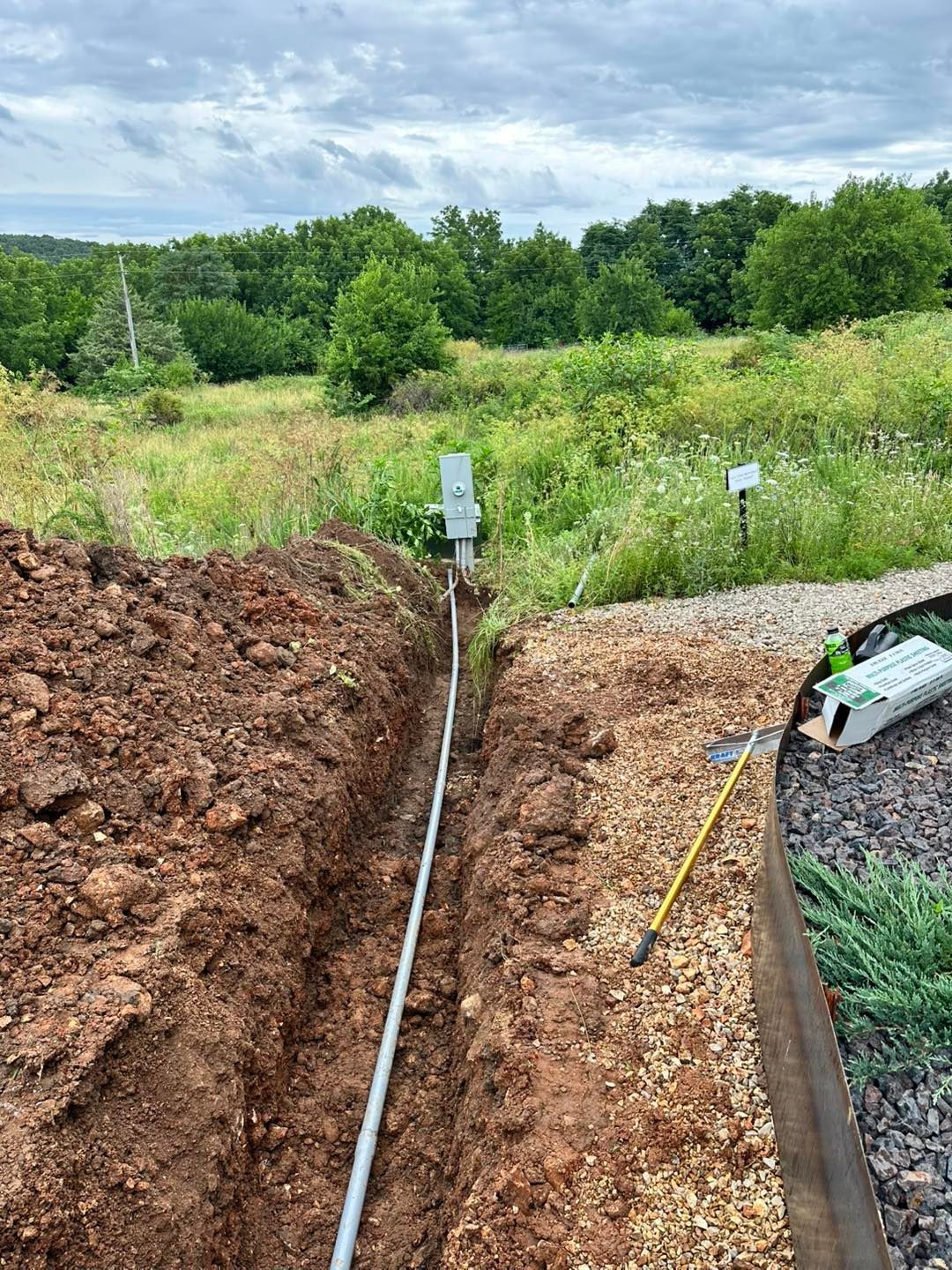 Trench with a laid pipe and markers in a grassy field under cloudy skies