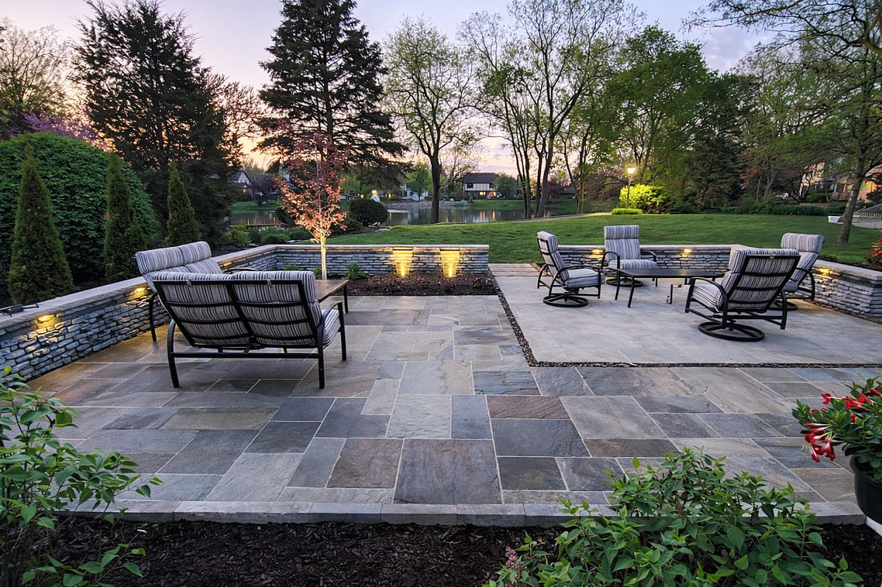 Stone patio seating area with black chairs overlooking a landscaped backyard and pond at sunset