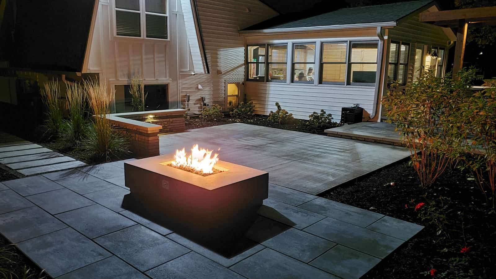 Lit fire pit on a patio at night, with a house and landscaping in the background.