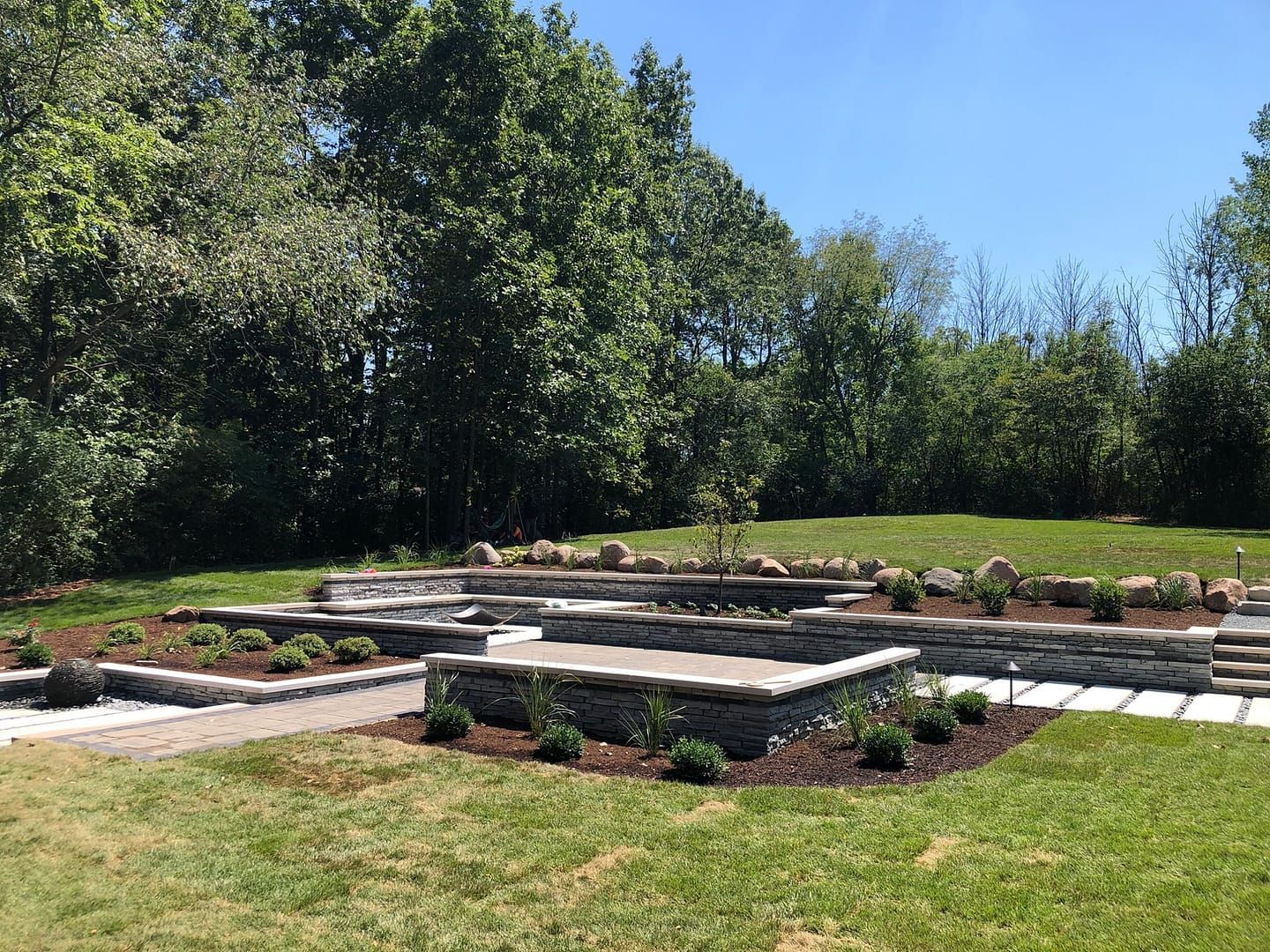 Raised stone garden beds in a sunny backyard with grass and trees beyond