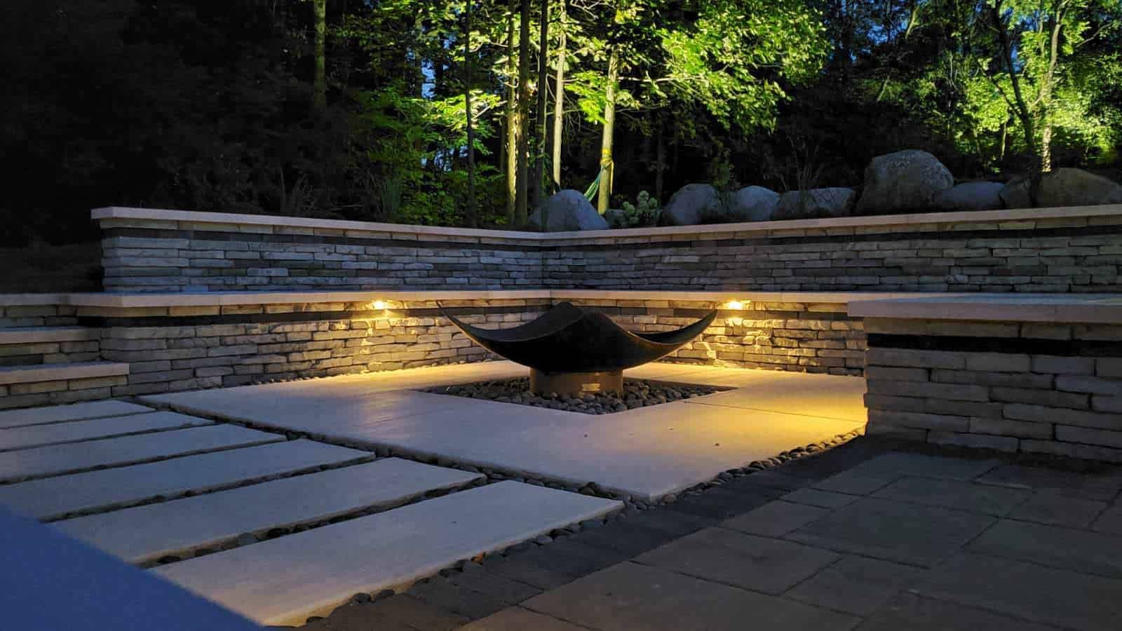 Nighttime view of a stone patio with a lit fire pit and surrounding stonework. Trees are illuminated in the background.