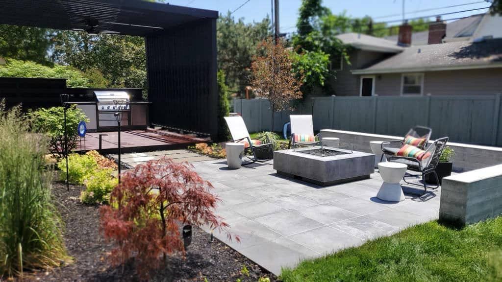 Backyard patio with grill area, fire pit, seating, and landscaping against a fence.
