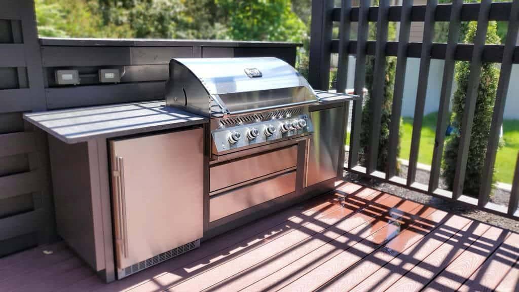 Stainless steel outdoor grill and refrigerator on a wooden deck, against a dark fence.