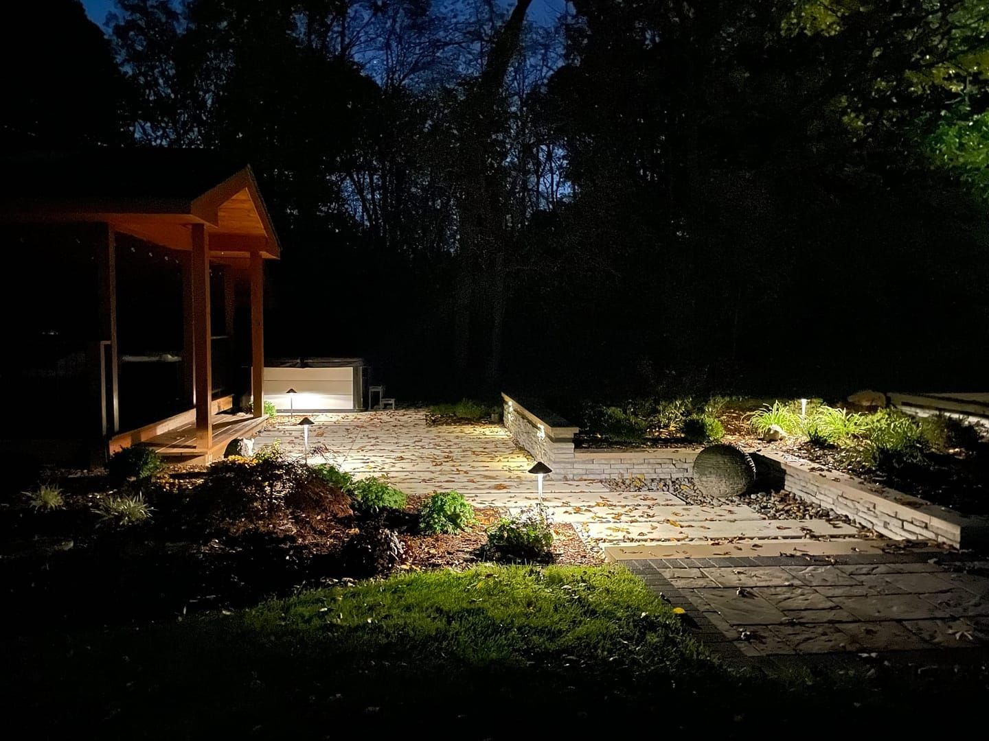 Nighttime garden path with stone walkway, wooden structure on the left, and bright lights illuminating plants.
