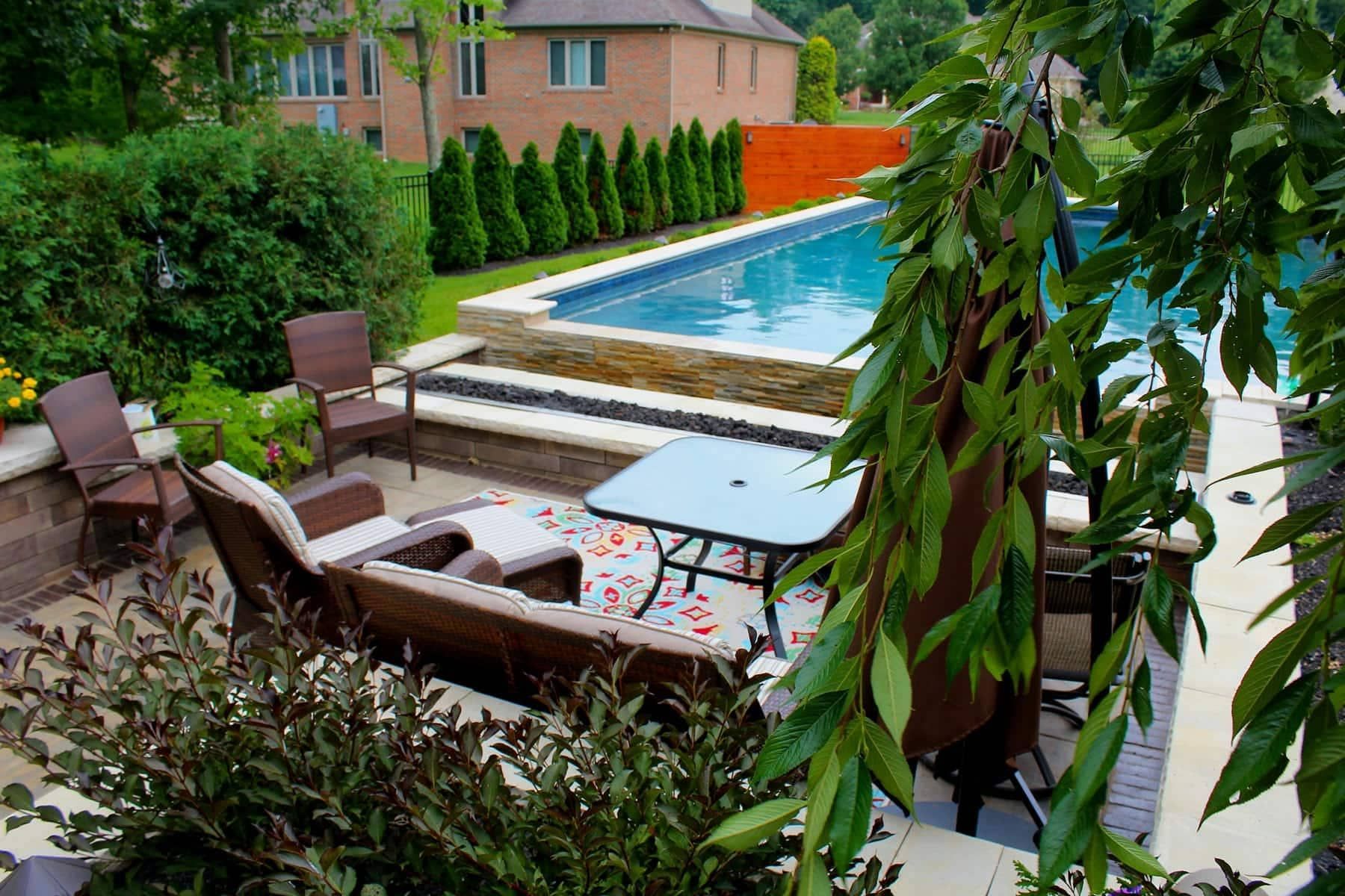 Patio with pool: lounge chairs, small table, green foliage, and a brick house in the background.