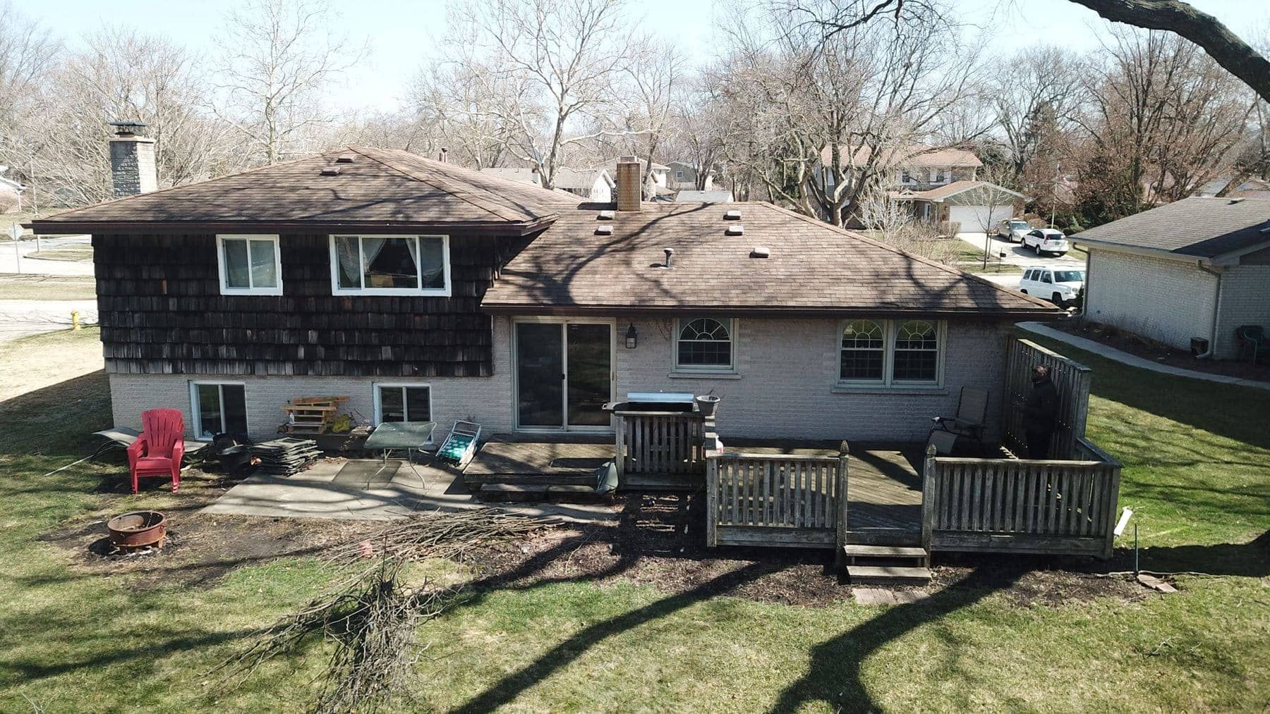 Backyard view of a two-story house with a wooden deck, brick patio, and bare trees in early spring.