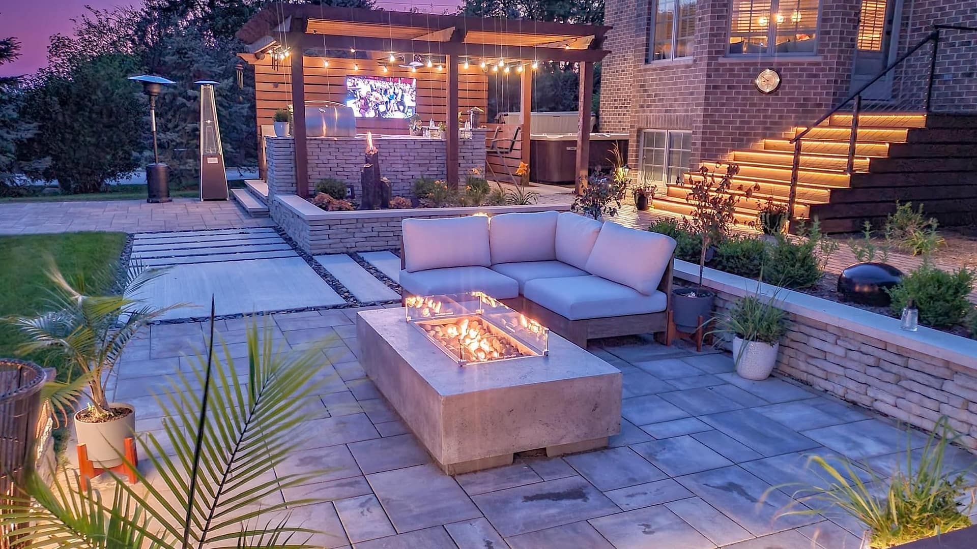 Outdoor patio with lit fire pit, sectional sofa, pergola bar, and steps at dusk.