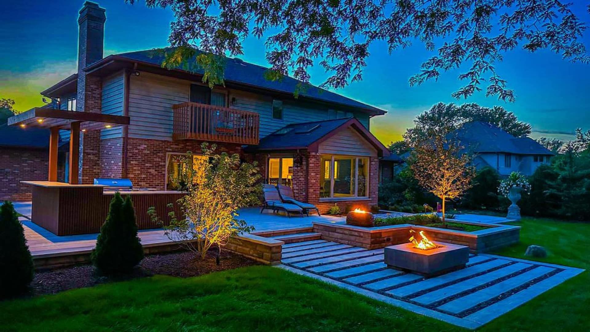 A cozy backyard at dusk with a brick house, patio, fire pit, and landscape lighting.