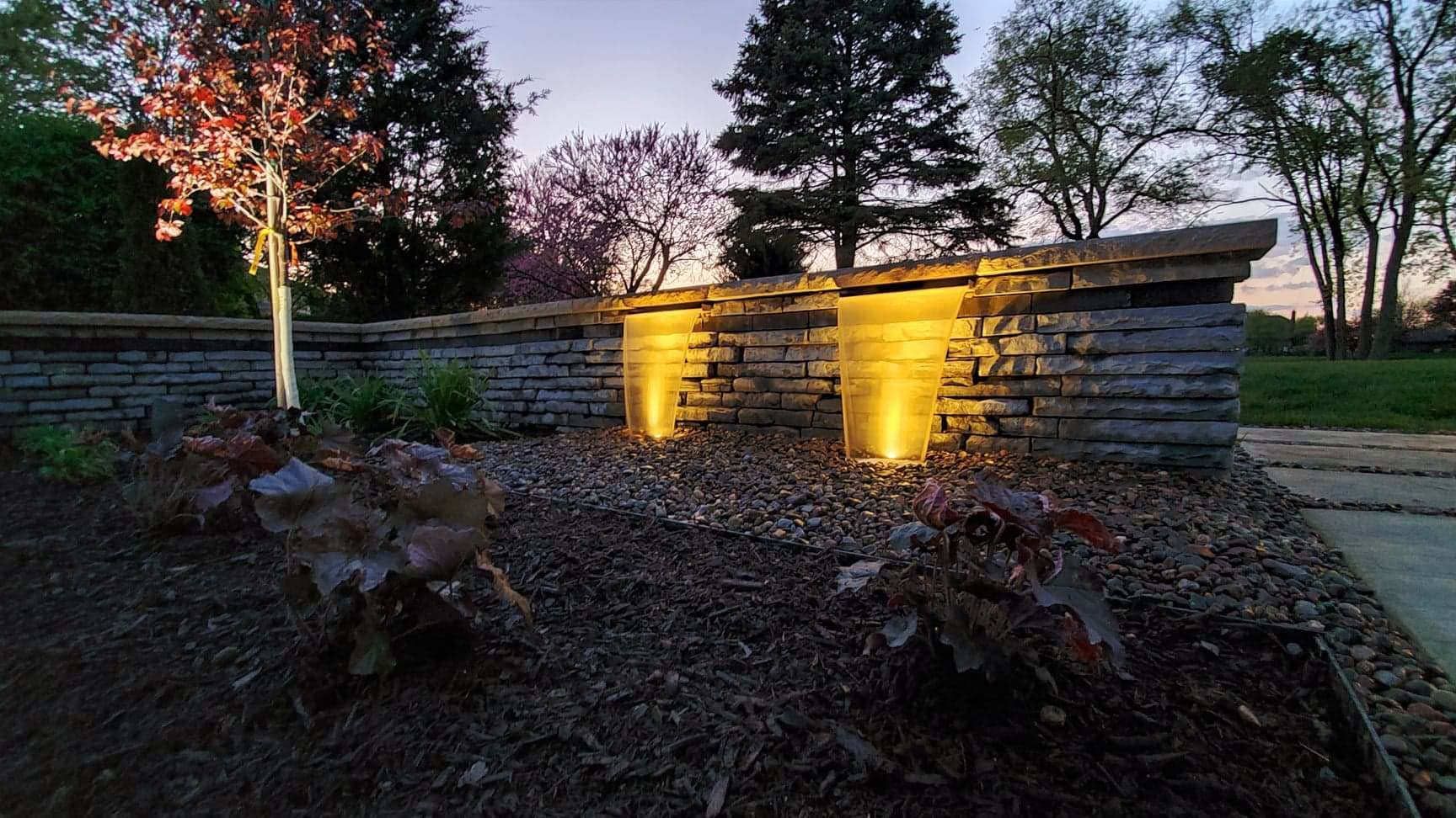 Stone wall with uplighting at dusk; landscaping in front.