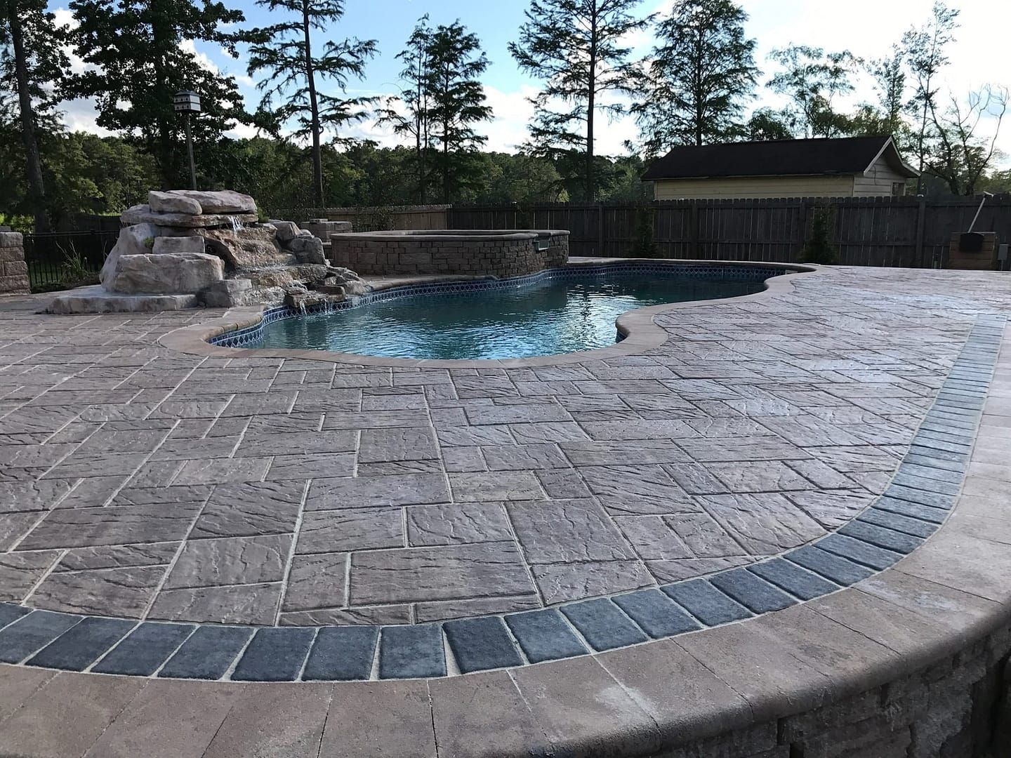 Pool with a stone patio, waterfall feature, surrounded by trees and a house.