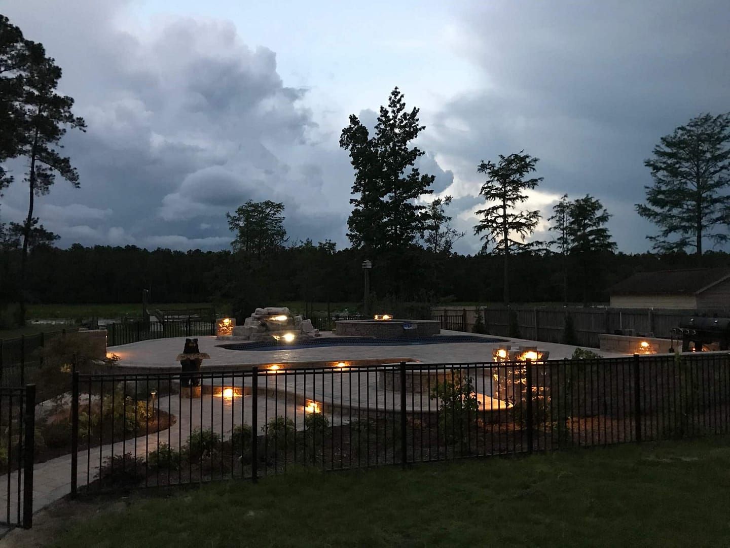 Evening view of a lit-up outdoor pool area with surrounding landscaping and a dark cloudy sky.