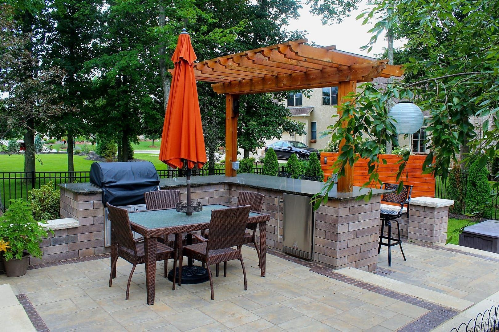 Outdoor patio with grill, dining set under orange umbrella and wooden pergola.