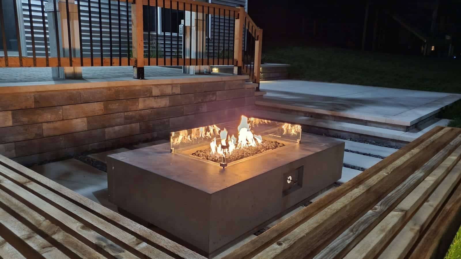 Fire pit with flames on a concrete patio at night, surrounded by wooden benches and a deck.
