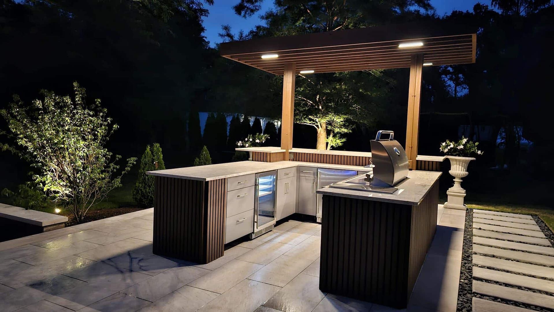 Outdoor kitchen at night with lights. Wooden bar, grill, fridge on a stone patio. Trees and bushes in the background.