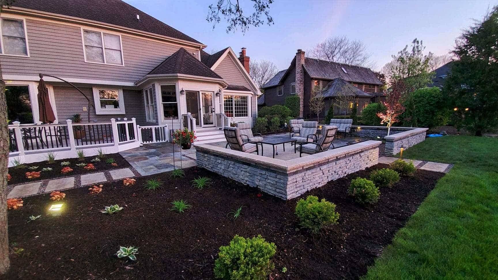 Backyard patio with fire pit and seating, adjacent to a house. Landscaped flowerbeds with new plants.