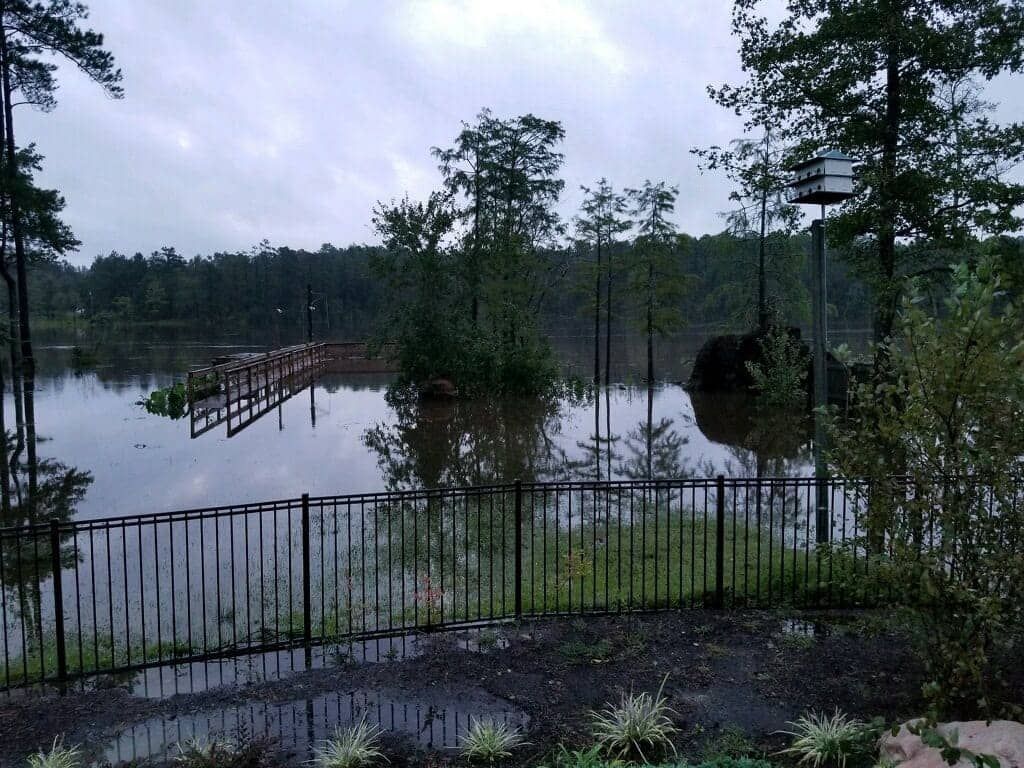 Flooded lakeside view with a partially submerged dock and fence. Overcast sky, dark water.