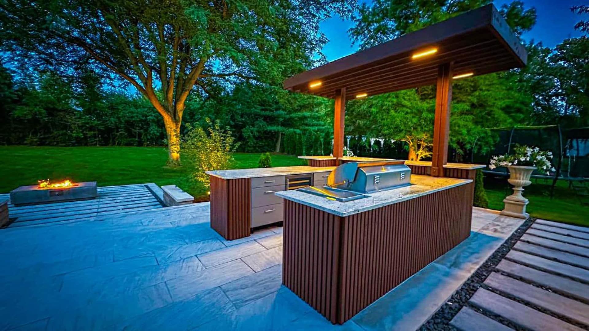 Outdoor kitchen with brown cabinetry, grill, and overhead lighting on a patio with a fire pit and lush lawn.
