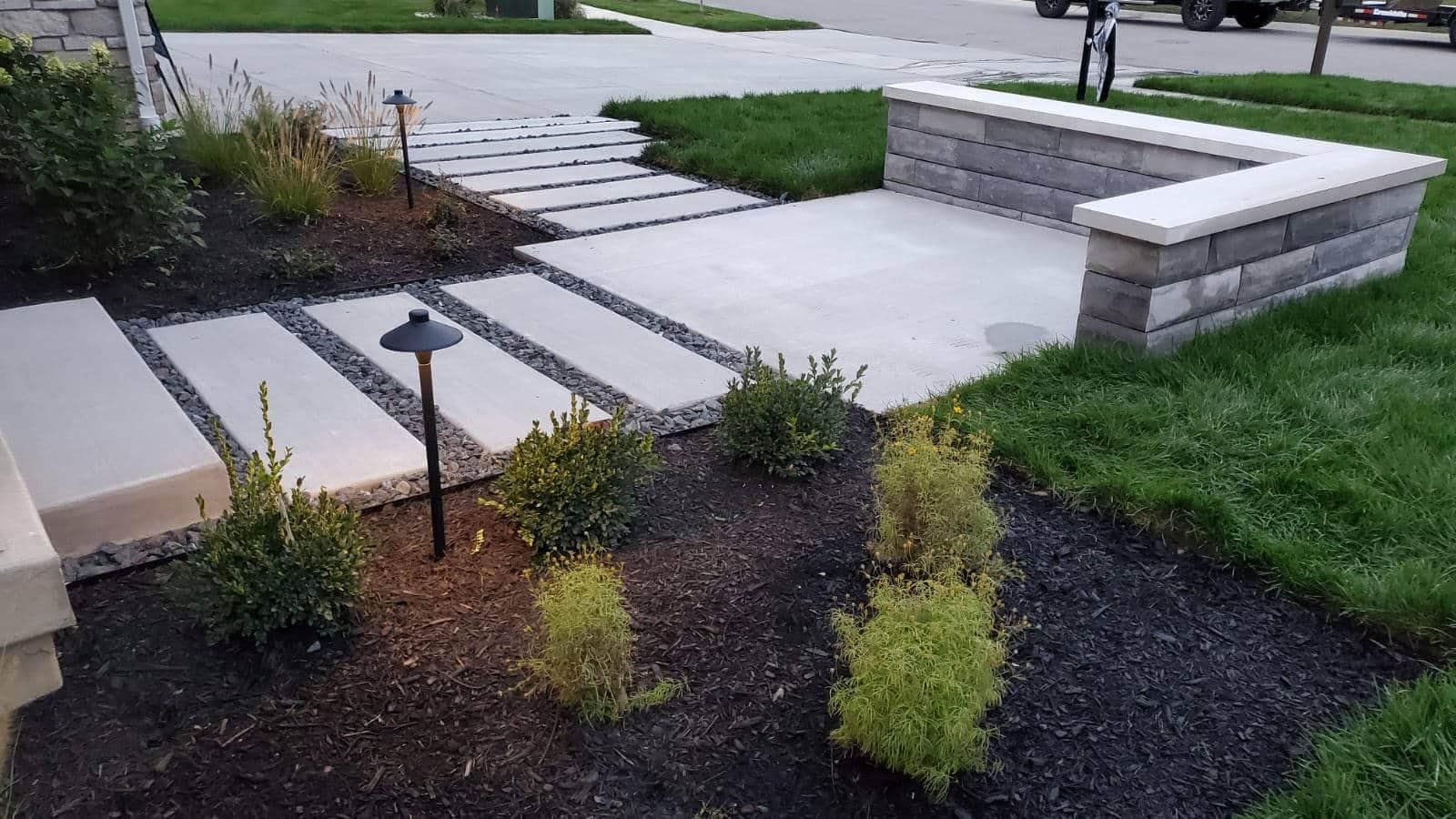 Stone walkway and small wall in a landscaped front yard with dark mulch and green plants.