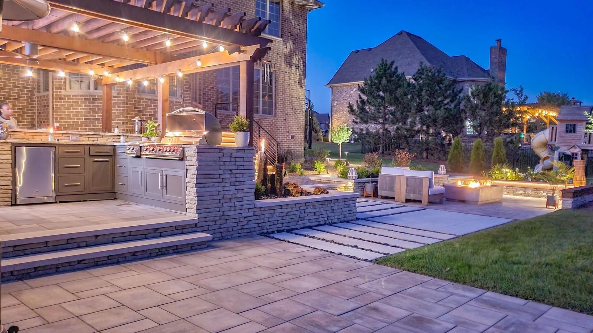 Outdoor patio with built-in grill, pergola lights, stone seating, and a tiled walkway at dusk
