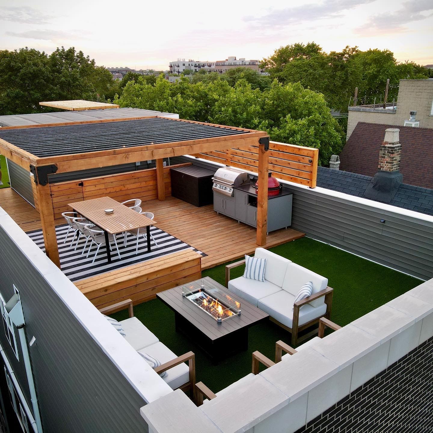 Rooftop deck with seating, pergola, grill, and fire pit. Green turf, wood decking, and light gray furniture.