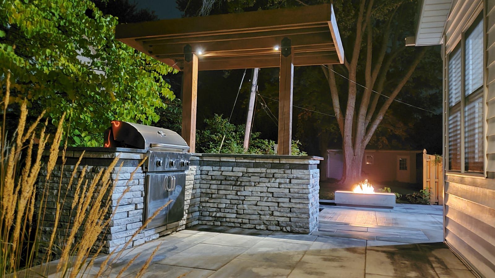 Outdoor patio with stone bar, grill, hot tub, and fire pit under a wooden pergola at night