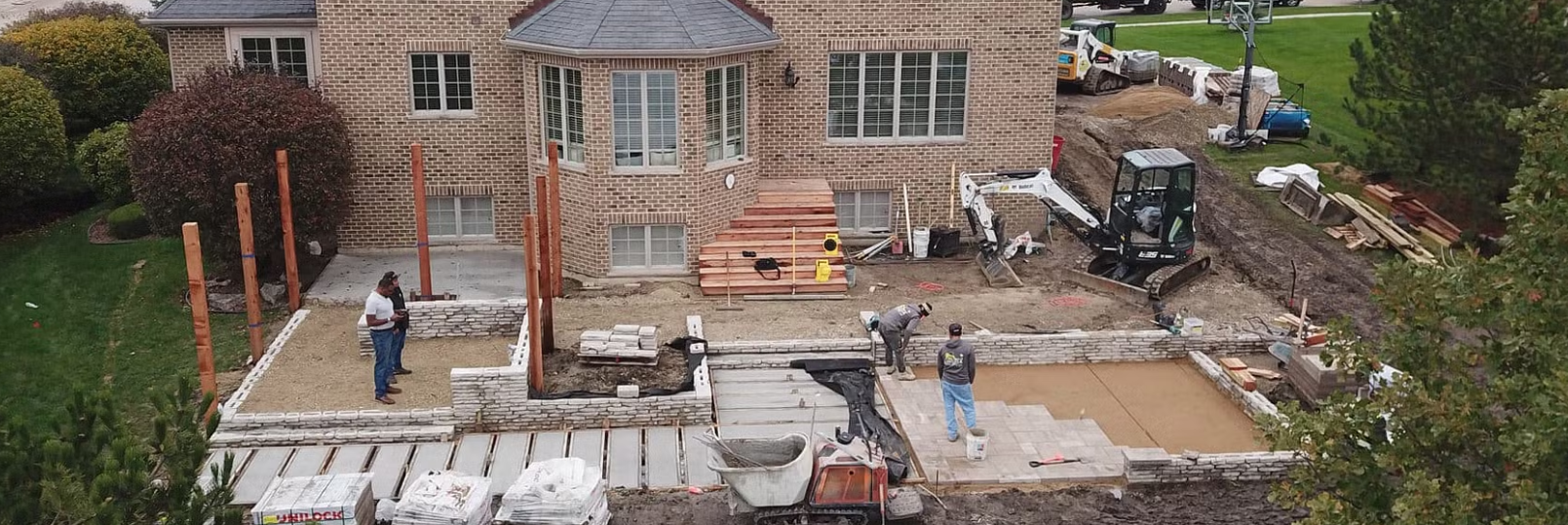 Construction workers building a patio at a house. A small excavator is visible.