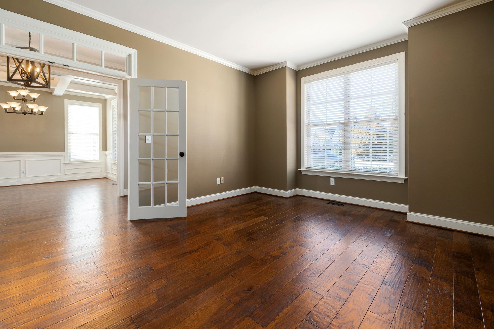 Empty living room with hardwood floors, tan walls, French doors, and a large window with blinds