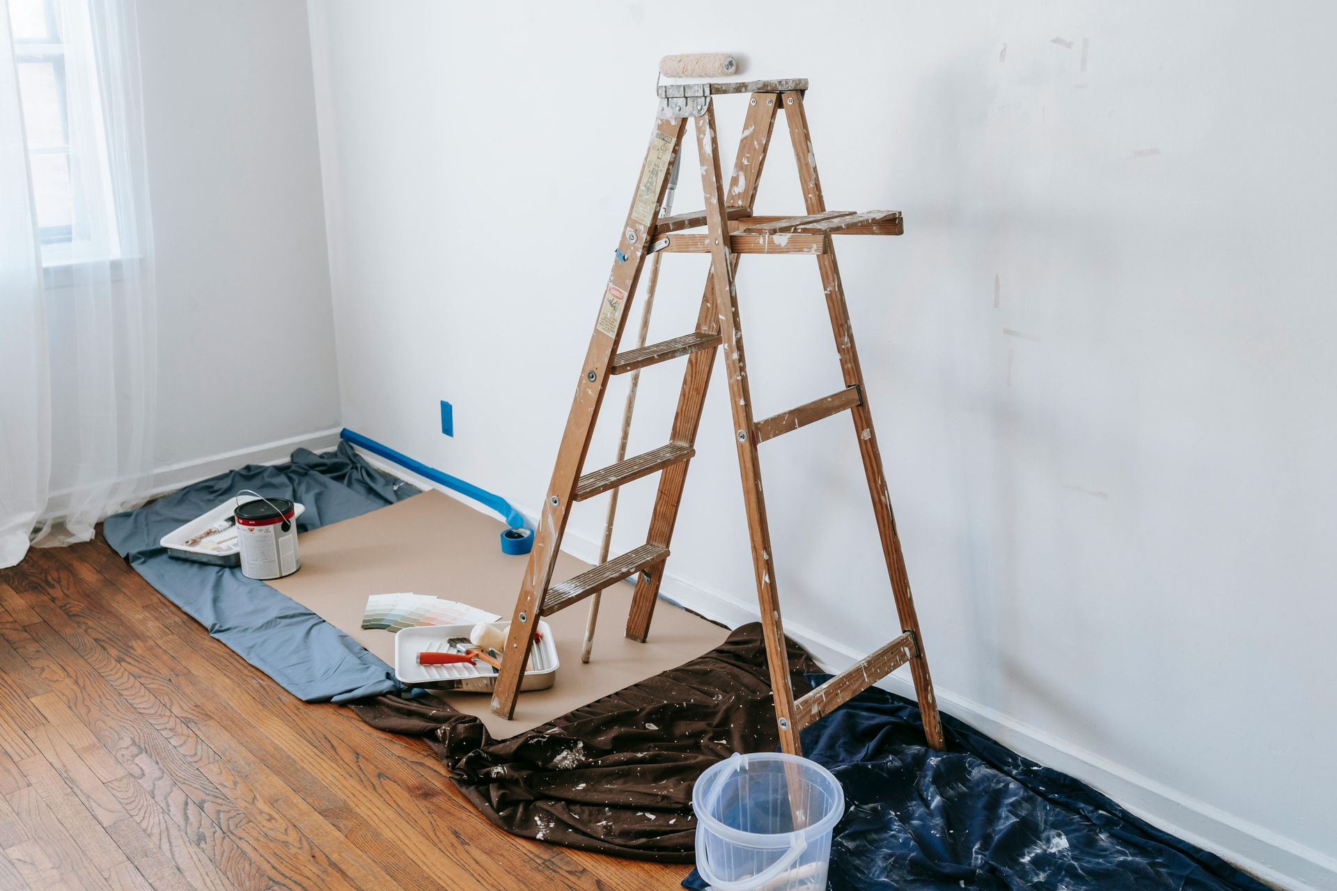 Ladder and painting supplies in a room with fresh white walls and covered hardwood floor