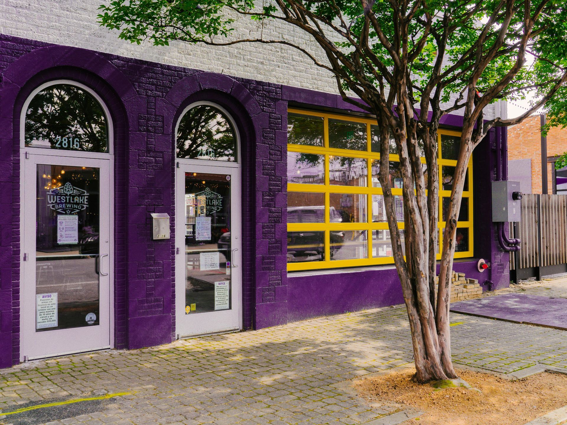 Purple storefront with arched windows and yellow garage-style door behind a tree on the sidewalk
