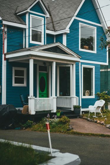 Turquoise two-story house with white trim and a small front porch beside a wet street.