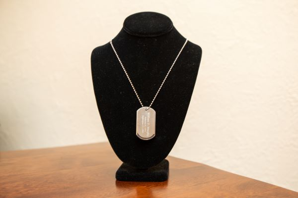 A silver dog tag necklace on a black velvet display bust, sitting on a wooden surface against a white wall.