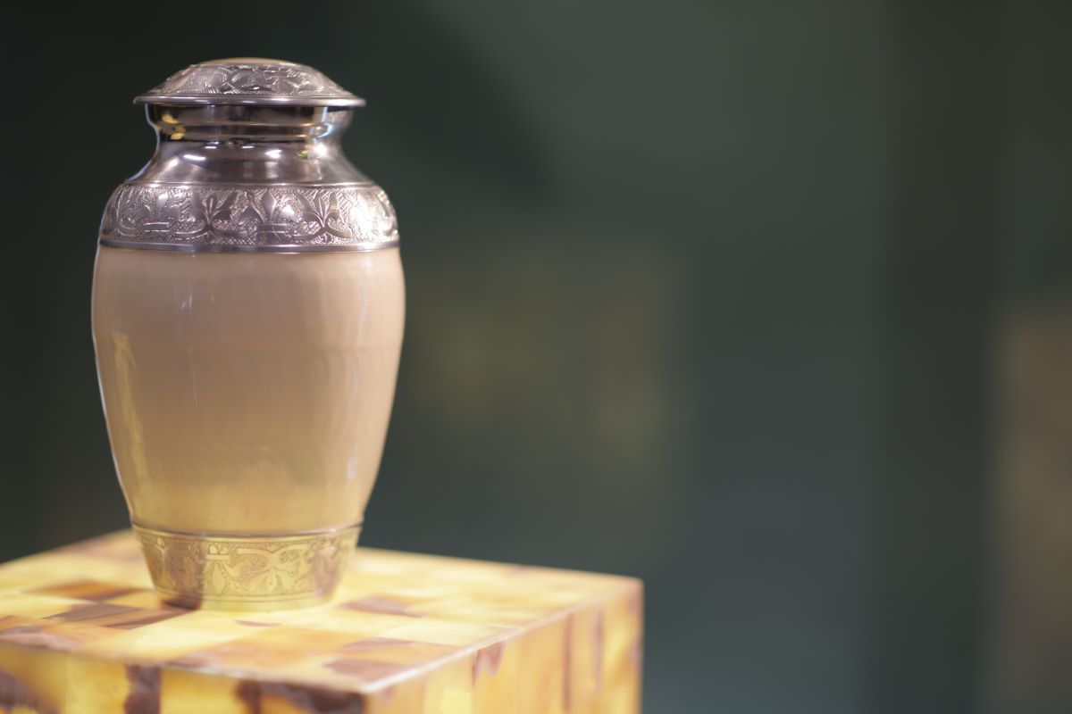 A small, ornate silver and tan urn rests on a patterned, square platform against a green background.