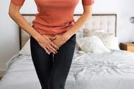 Woman in orange top and black pants, holding hands over her crotch in a bedroom.