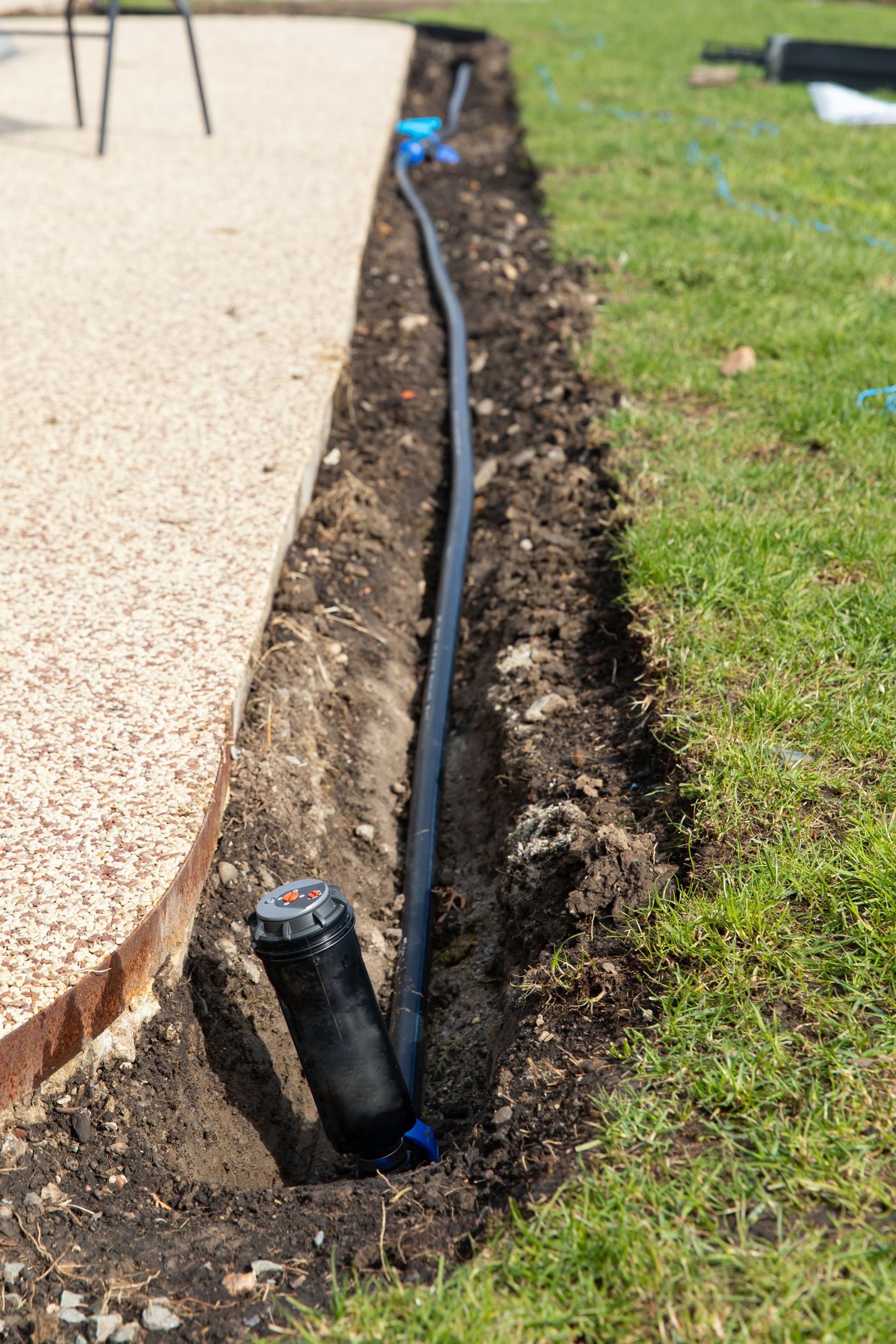 A sprinkler head installed in a narrow trench next to a paved patio, with black tubing running along the ground.