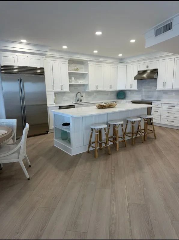 Bright white modern kitchen with island, stainless fridge, and four bar stools on wood flooring