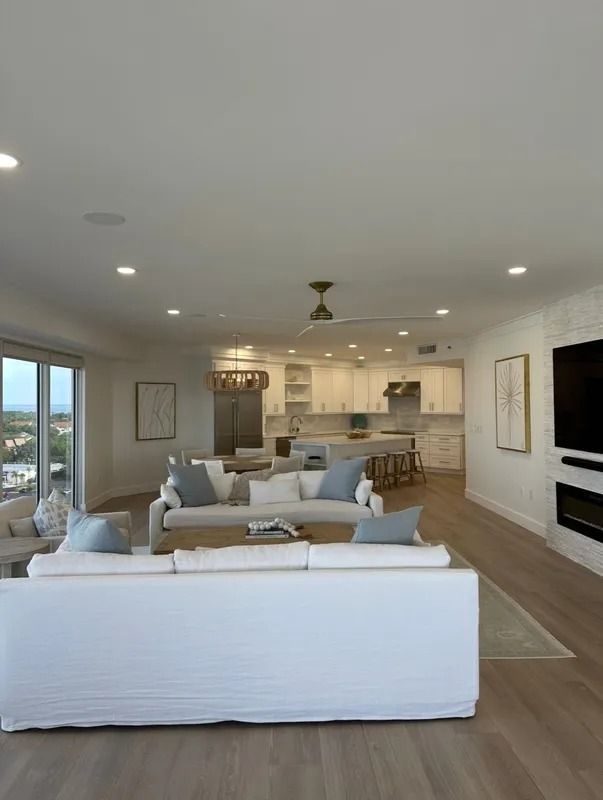 Bright open-concept living room with white sofas, kitchen in back, and large windows overlooking the view