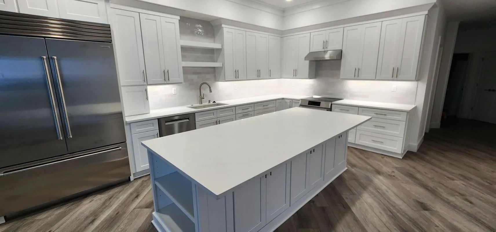 Bright modern kitchen with white cabinets, stainless steel refrigerator, and large white island with dark wood flooring