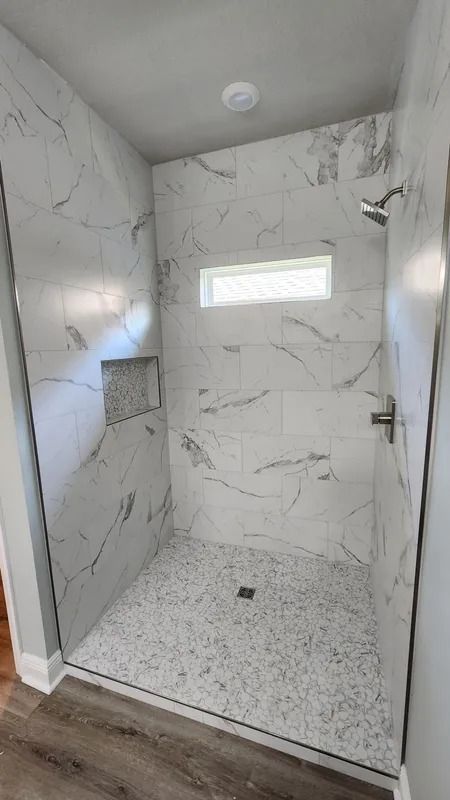 Modern tiled walk-in shower with glass door, white marble-look walls, pebble floor, and small horizontal window
