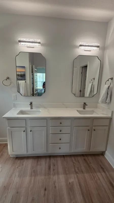 Bright bathroom vanity with double sinks, white cabinets, mirrors, and wall lights above a wood floor.