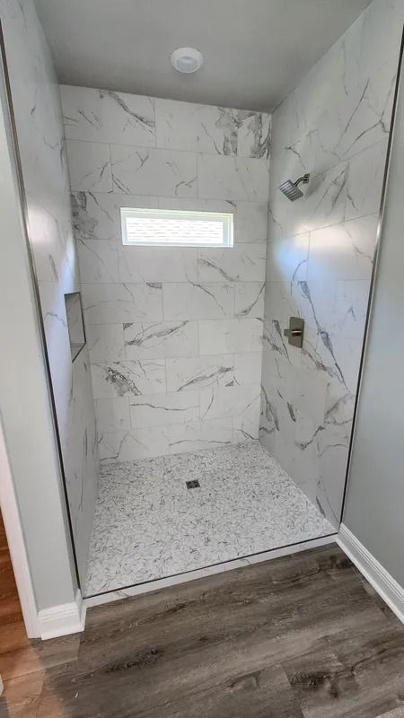 Empty tiled shower with marble-look walls, white pebble floor, and a small horizontal window.