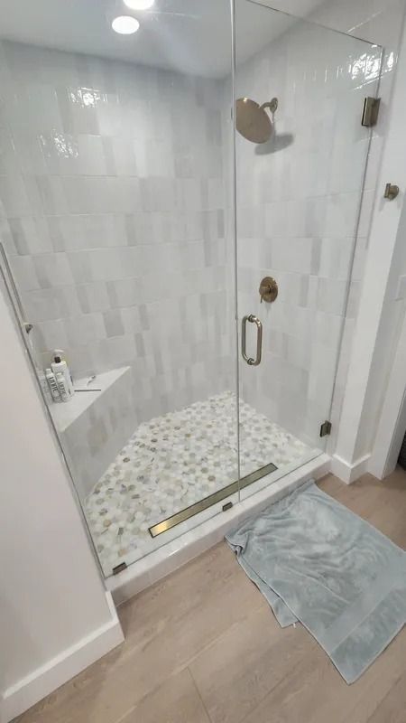 Glass shower stall with white tile walls and pebble floor in a bathroom, beside a blue bath mat