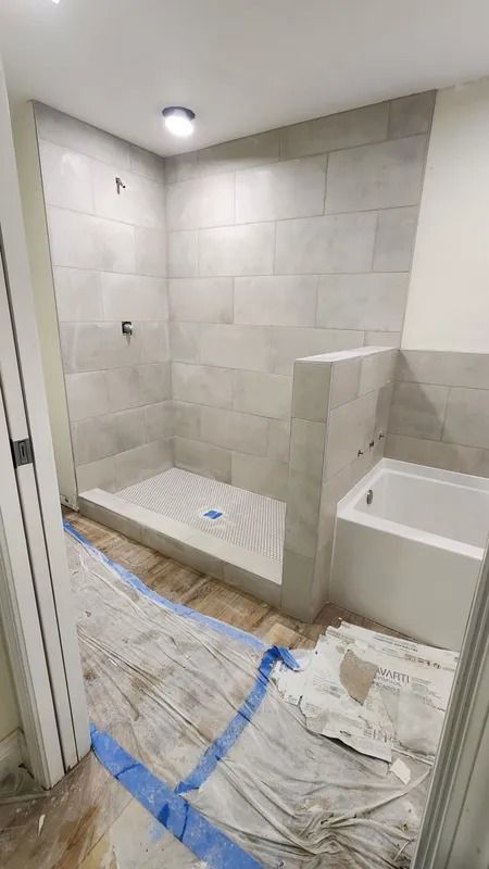 Tiled bathroom under renovation with shower bench, bathtub, and plastic-covered floor.